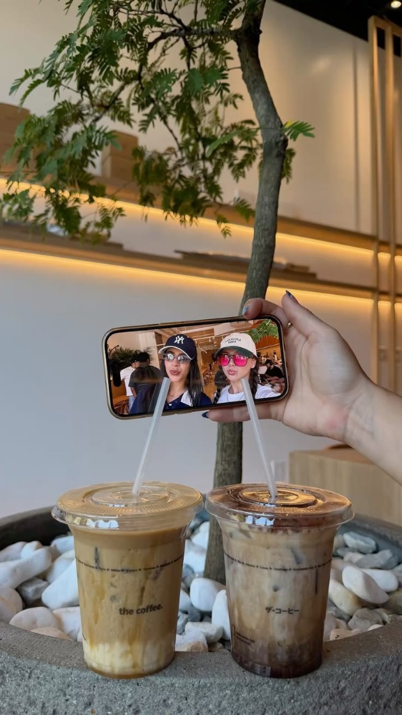 Trend Đồ Uống Cho Gen Z: Quán F&B Nên Theo Điều Gì, Bỏ Điều Gì? 6 Khách hàng cầm điện thoại selfie check-in bên hai ly cà phê đá đặt trên chậu cây sỏi trắng tại quán cafe