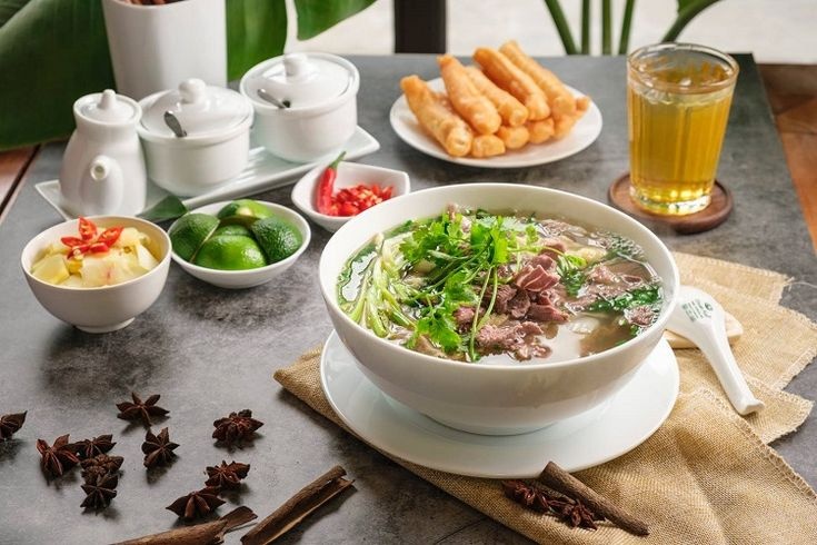 20 Món Ngon Hà Nội Nên Thử Khi Khám Phá Ẩm Thực Thủ Đô 5 Bát phở bò tái Hà Nội nóng hổi rắc nhiều hành ngò bên cạnh đĩa quẩy giòn, tỏi giấm, chanh ớt và ly trà đá