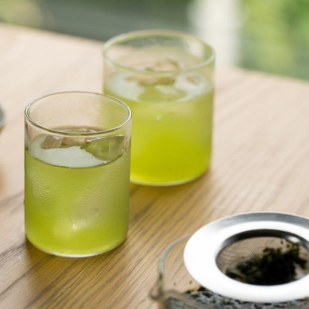 Cold Brew Tea Là Gì? Có Phù Hợp Với Menu Quán F&B Không? 5 Hai ly trà xanh Cold Brew ủ lạnh màu xanh cốm kèm đá viên đặt cạnh bình lọc trà inox trên bàn gỗ quán cafe